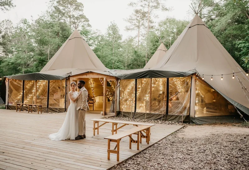 Bride and groom standing on the dancefloor A couple wearing wedding attire stands on a wooden deck in front of a large tent decorated with string lights in a wooded area.