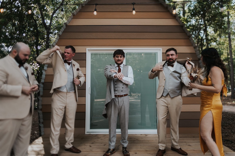 A groom and his groomsmen, all in beige suits, get ready outside a cabin. One woman in a yellow dress stands nearby holding a bouquet.
