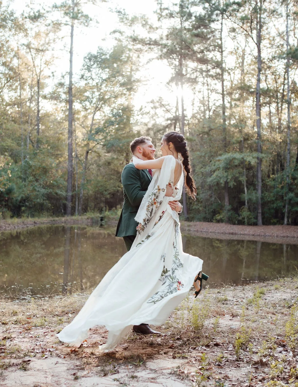 A wedding couple moment in nature among the trees A couple embraces near a pond in a forest setting. The woman wears a flowing white dress with floral designs, and the man is in a green suit. Sunlight filters through the trees.