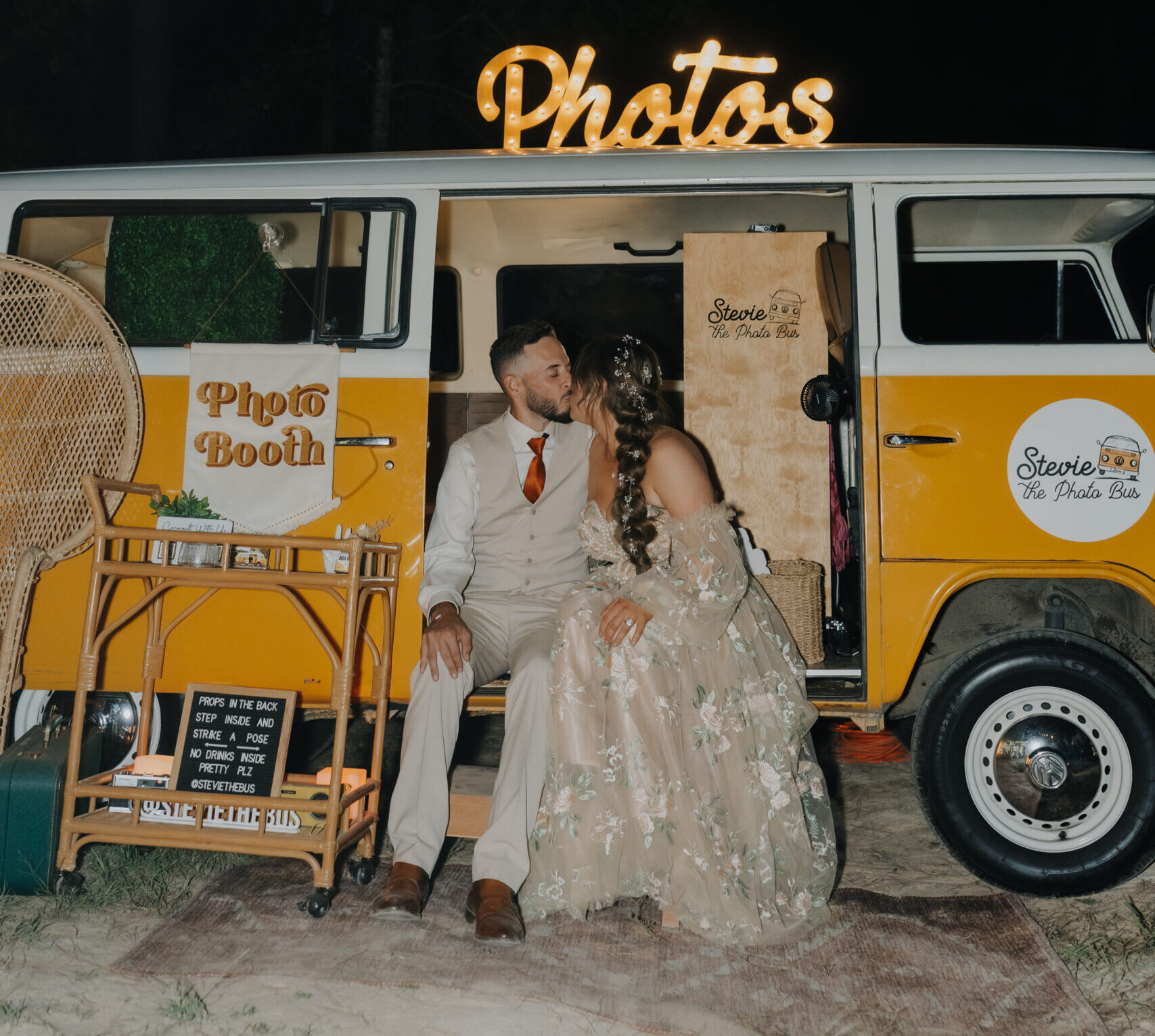 A couple sits in front of a vintage yellow van with a "Photos" sign. The van is set up as a photo booth at night. The man is in a light suit, and the woman wears a floral dress.