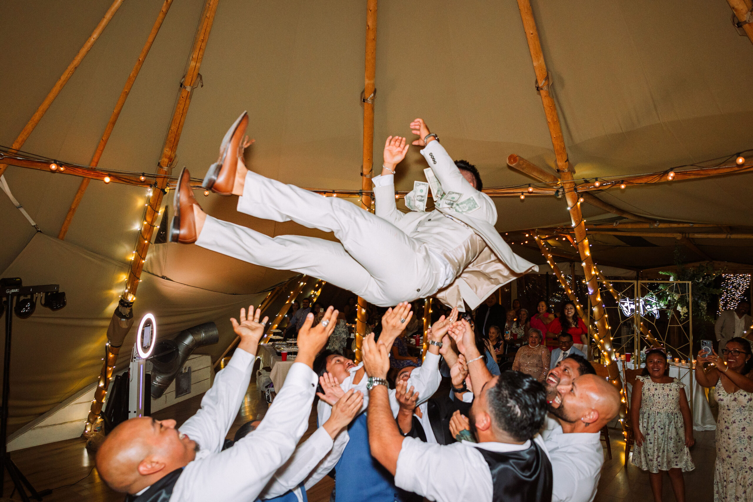 Group of people toss a person in a white suit into the air inside a decorated tent with string lights.