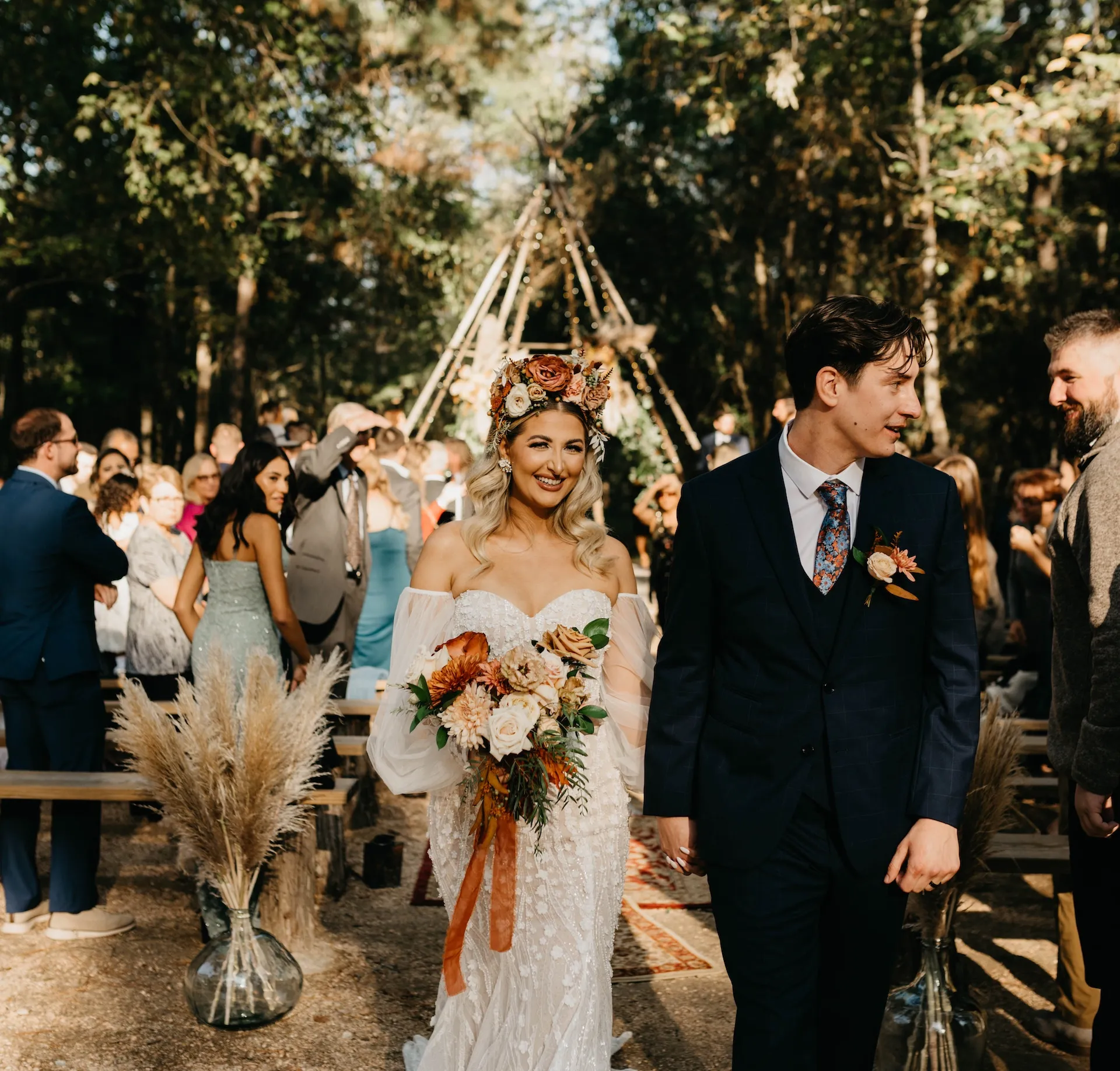 Bride and groom walking hand in hand, surrounded by guests in a forested outdoor wedding setting, with a floral bouquet and decorative tipi in the background, highlighting At The Shire's enchanting atmosphere for year-round celebrations.