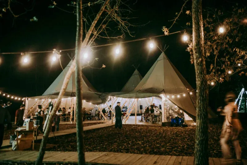 Nordic tipis illuminated by string lights at night, showcasing a festive outdoor wedding setting with guests mingling, emphasizing weather-resistant design for events at At The Shire in Huntsville, Texas.