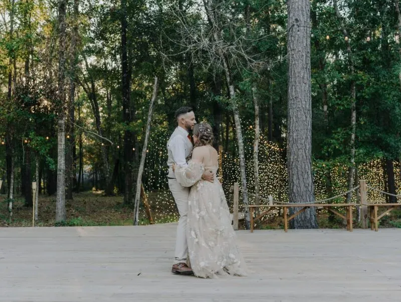 Couple dancing on an outdoor wooden platform surrounded by trees and twinkling lights, showcasing a romantic setting for an outdoor wedding at At The Shire in Huntsville, Texas.