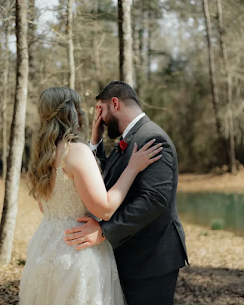 A bride and groom stand outdoors by a pond, facing each other. The groom covers his face with one hand while the bride rests her hand on his shoulder.