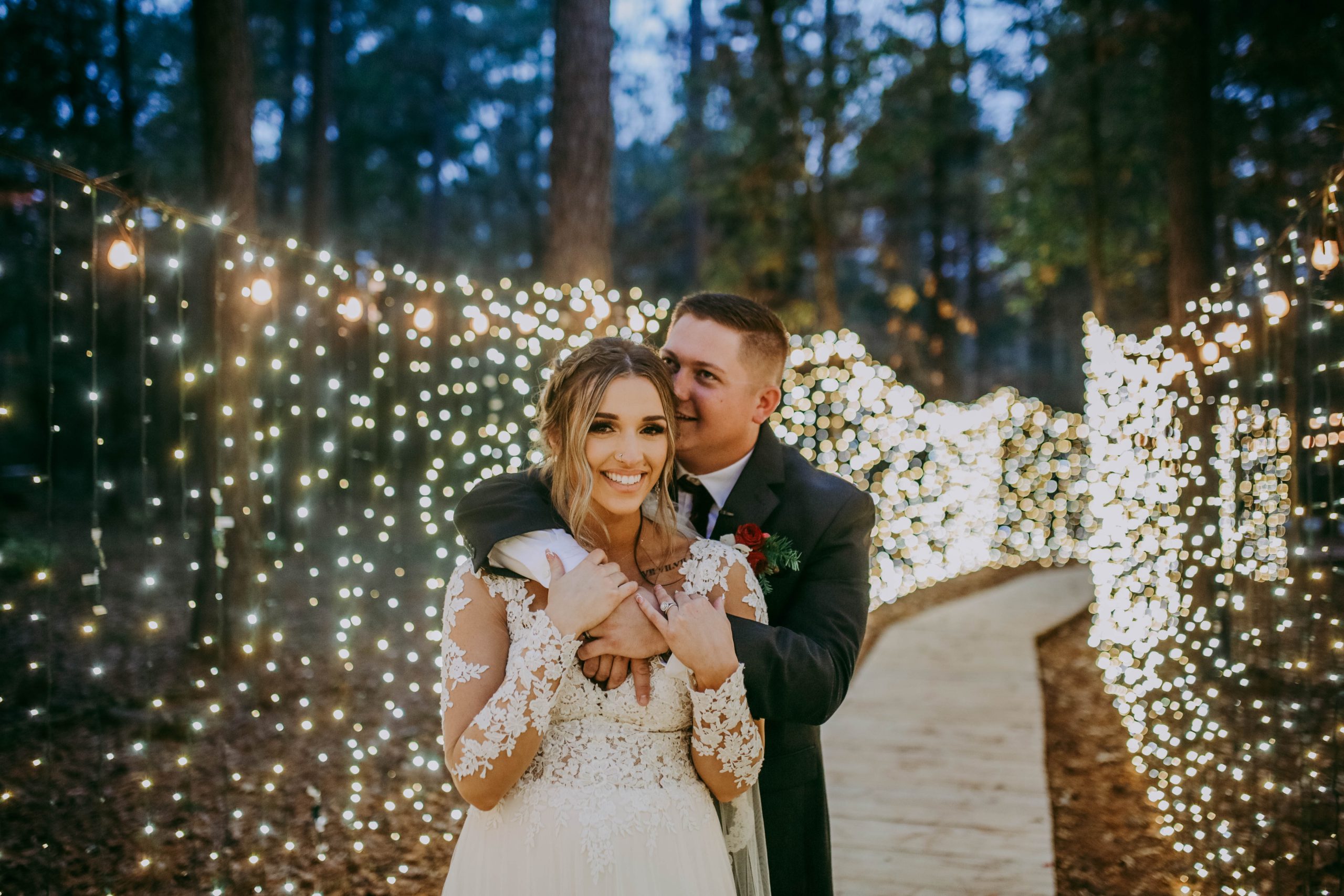 A bride and groom stand on a wooded path decorated with string lights; the groom hugs the bride from behind, both smiling.